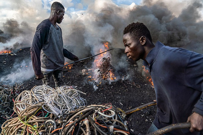 A young man is pictured burning electrical wires to recover copper at Agbogbloshie, Ghana, as another metal scrap worker arrives with more wires to be burned. (Image by Muntaka Chasant, published under a CC-BY-SA-4.0 license.)